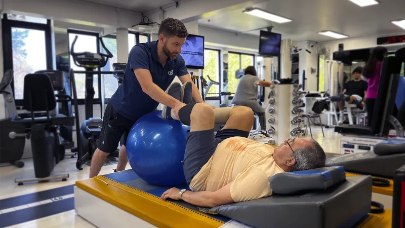 Kinesiólogo asistiendo a paciente adulto mayor recostado en colchoneta mientras realiza ejercicio con balón terapéutico en KinesicWork