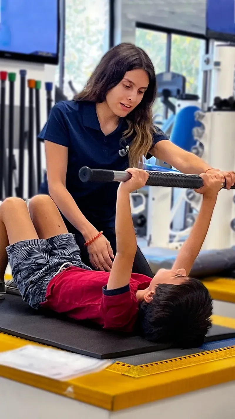 Kinesióloga asistiendo a niño en ejercicio terapéutico con barra liviana durante sesión de rehabilitación musculoesquelética